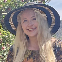 Headshot of a smiling woman wearing a wide-brimmed hat, with long blonde hair, against trees in the background.