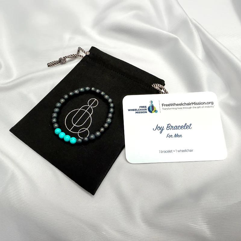 A men's Joy bracelet made of black beads, with four teal colored beads, with a black pouch bearing the Free Wheelchair Mission logo.